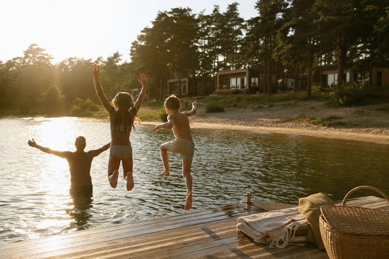 Kinder springen in der Abendsonne von einem Steg in einen Badesee, dahinter Ferienhäuser zwischen Kiefern