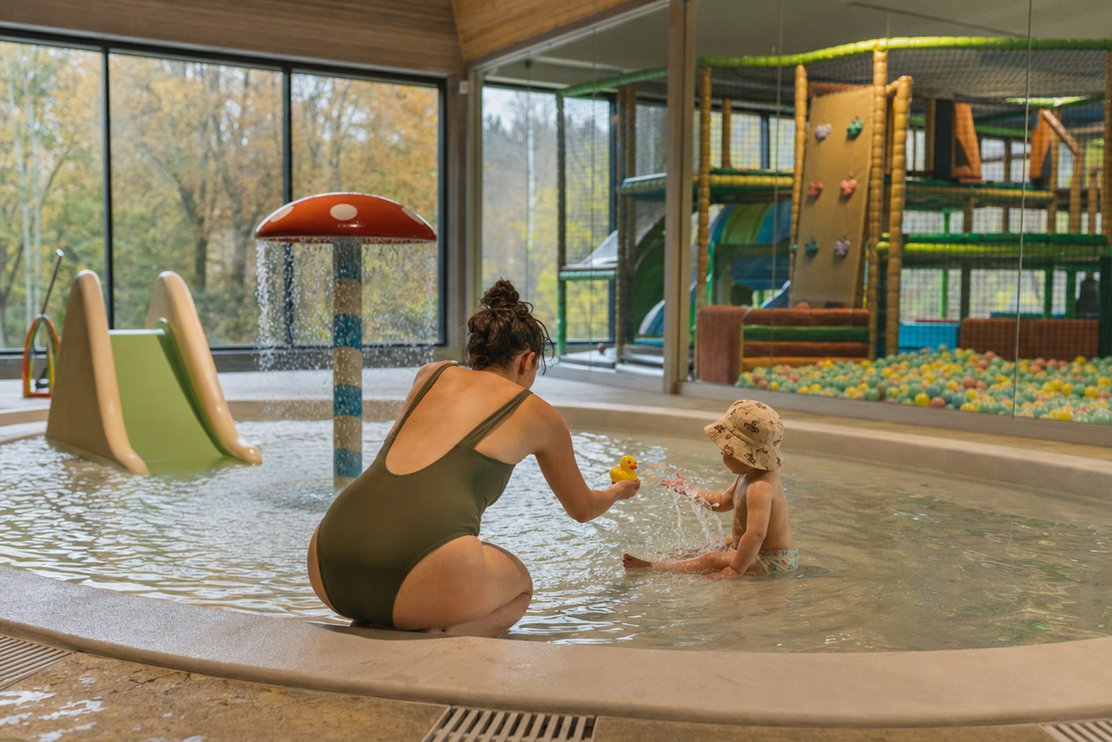 Mutter kniet am Rand eines flachen Kinderplanschbeckens im Hallenbad und reicht ihrem Kleinkind mit Sonnenhut eine Gummiente, daneben ein Pilz-Wasserspiel und eine kleine Rutsche, hinter einer Glaswand ein Indoor-Spielplatz mit Bällebad